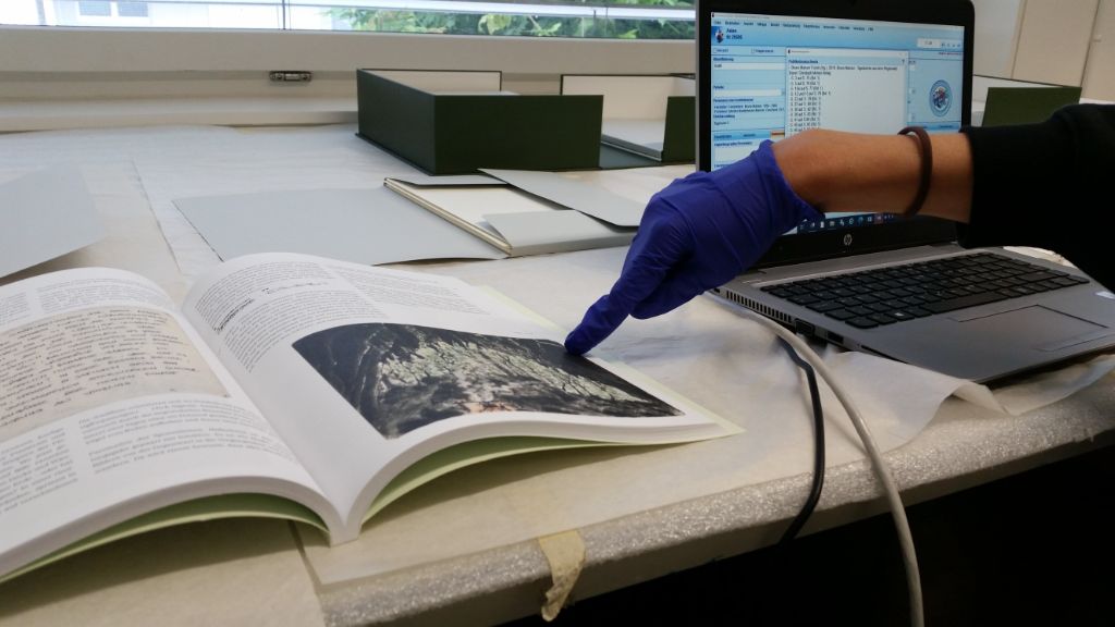 Auf einem Tisch liegt ein aufgeschlagenes Buch mit Text und Bildern. Eine Hand in blauem Handschuh zeigt auf eines der Bilder. Dahinter steht ein Laptop.