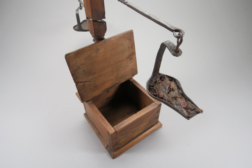 Holzbox mit offenem Deckel und darüber hängt ein flaches Eisengefäss mit Talgresten drin