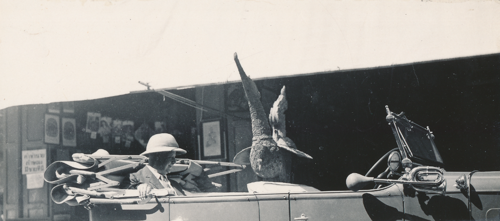 Schwarz-Weiss-Foto, in den Zentrum ein Oldmobil steht mit Hupe. Im Auto sitzt ein Mann im Anzug mit Tropenhut, vor ihm steht eine Vogelfigur.