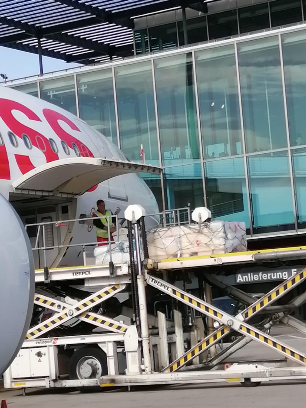 Vor einem Glasgebäude an einem Gate sieht man einen kleinen Teil eines Swiss-Flugzeugs, in dessen Bauch Kisten verladen werden mit Hebebühnen. 