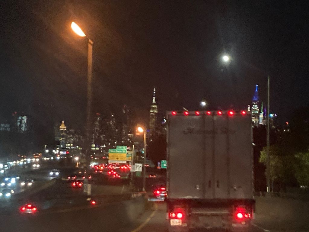 Blick aus einem Auto auf die Fahrzeuge davor, darunter eine Lastwagen, und auf den Gegenfahrbahnen.  Es ist Nacht und überall brennen Lampen. Im Hintergrund sieht man die Silhouette von New York mit beleuchteten Wolkenkratzern.