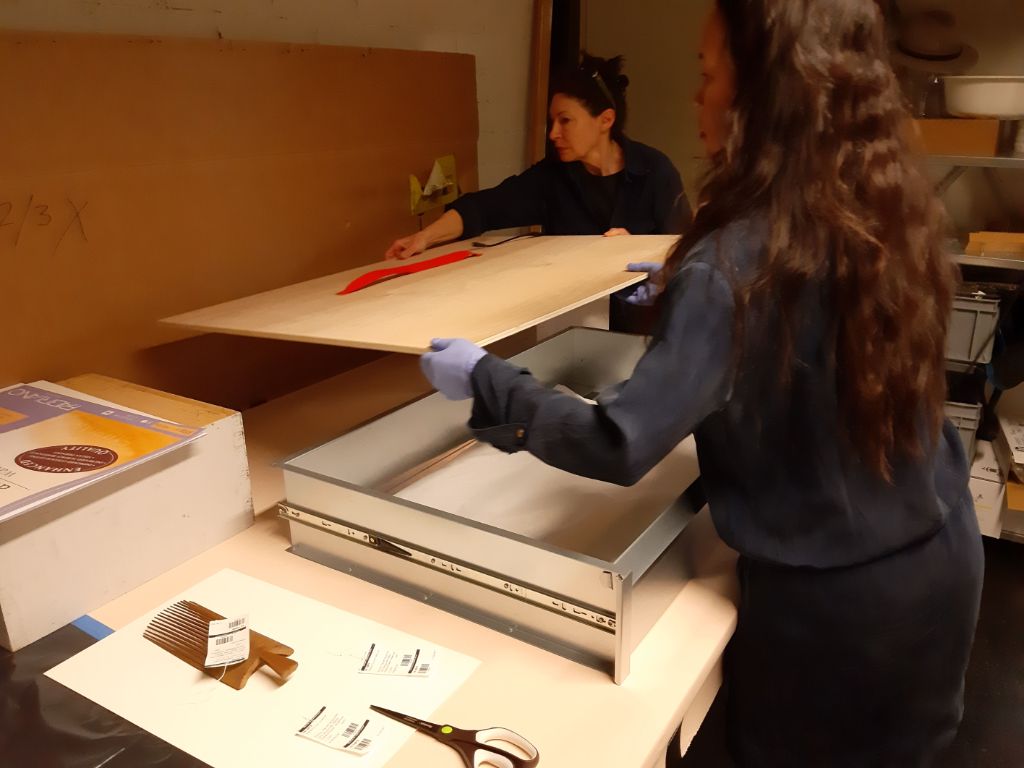 Zwei Frauen heben eine Holzplatte hoch, um sie auf einen grauen Behälter zu legen. Im Vordergrund liegen Papiere und eine Schere.