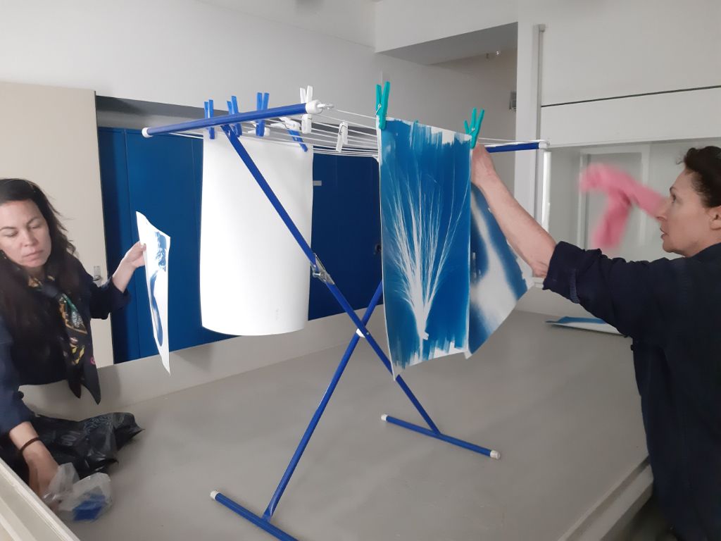 Zwei Frauen stehen neben einem blauen Wäscheständer, an dem schon zwei Blätter hängen. Sie befestigen zwei weitere Blätter daran mit Wäscheklammern.