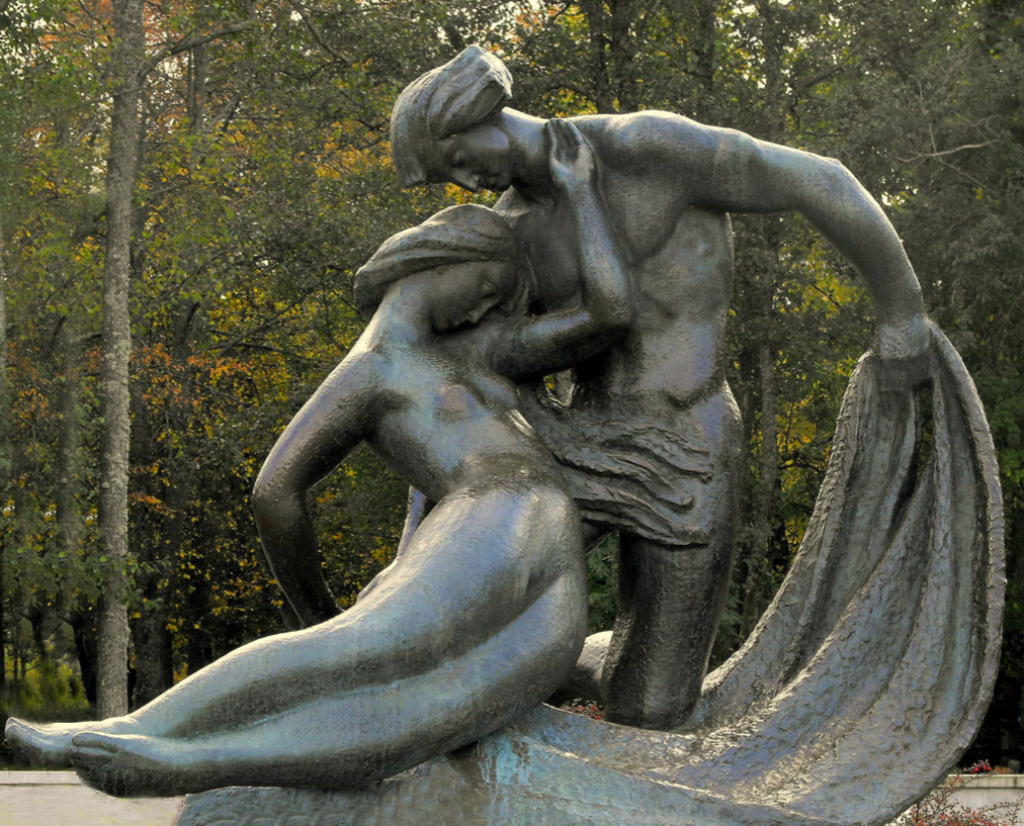 Skulptur vor Bäumen. Ein Mann kniet auf einem Tuch, das er in der linken Hand hält. An ihn lehnt sich eine Frau mit langen Haaren.