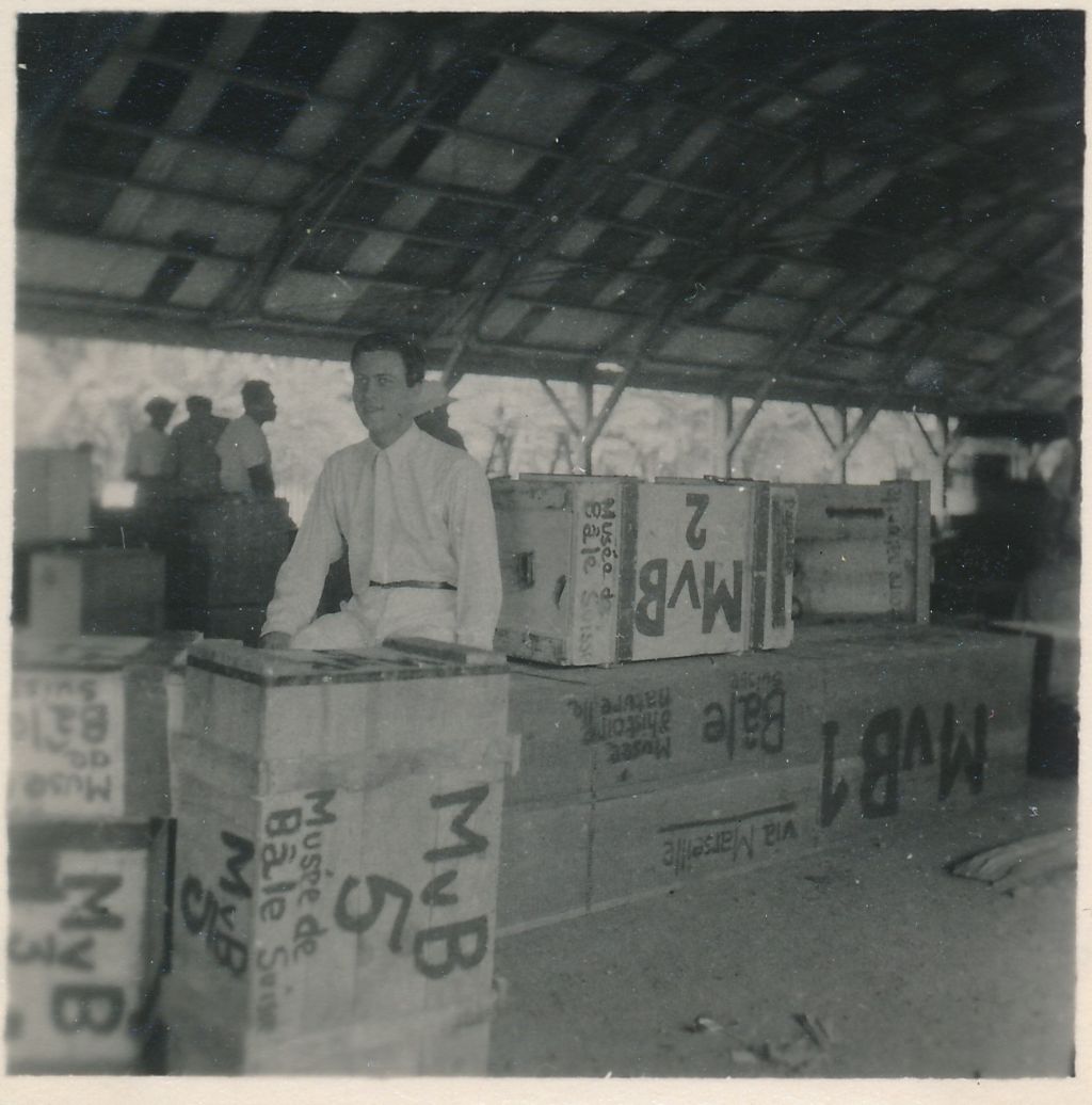 Auf einem viereckigen Schwarz-Weiss-Foto sitzt ein weissgekleideter Mann mit dunklem Haar auf einer Kiste, umgeben von vielen weiteren Holzkisten. Über ihm ist ein Holzdach zu sehen und im Hintergrund weitere Menschen.