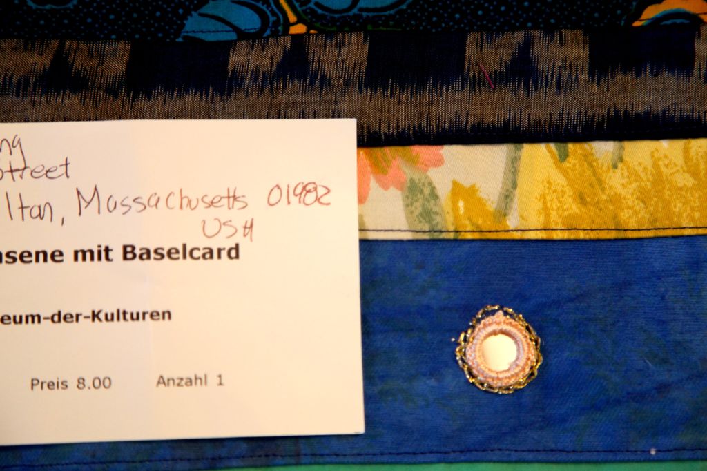 Ausschnitt einer Tasche aus blauem, geblümten und dunkelbraunem Stoff. Auf der Tasche liegt eine weisse Karte, auf der ein Name und eine Adresse von Hand notiert ist.