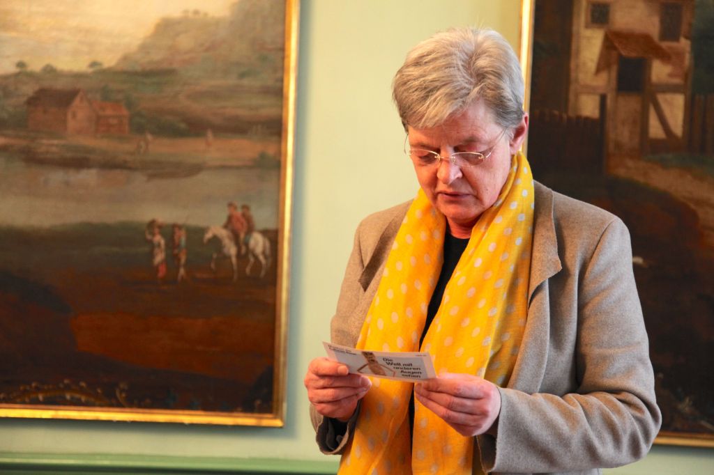 Eine Frau mit kurzem grauem Haar, Brille, gelbem Schal und grauer Jacke steht vor einer grünen Wand mit zwei Bildern und hält eine Karte in der Hand. Sie liest, was auf der Karte steht.