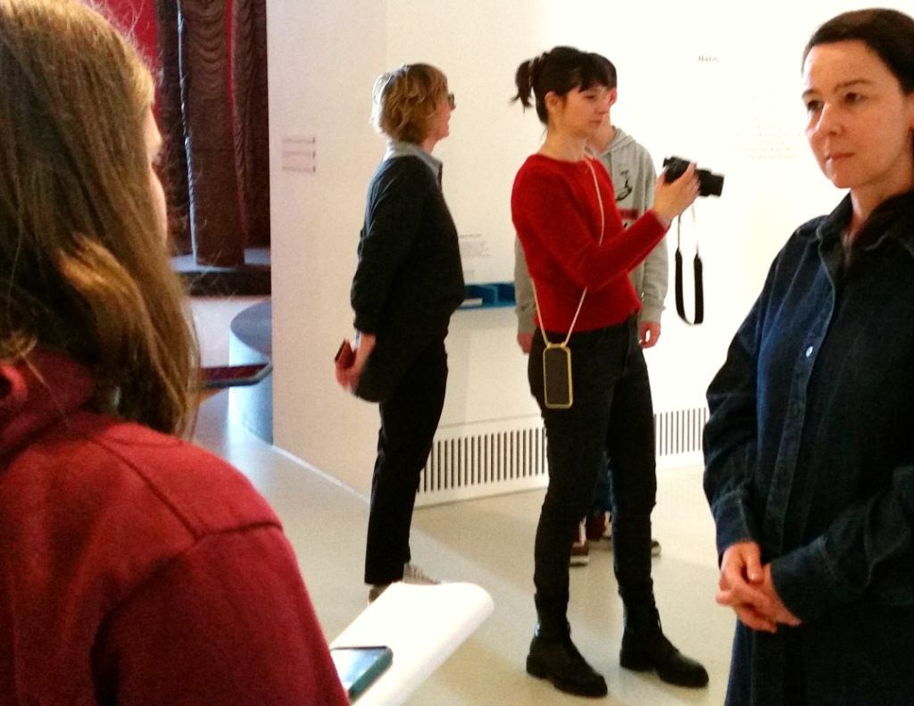 In einem hellen Ausstellungsraum stehen fünf Menschen. Vorne links eine junge Frau mit langem braunen Haar, sie hält einen Block und ein Handy in der Hand. Rechts von ihr schaut sie eine dunkel gekleidete Frau mit braunem Haar an. Im Hintergrund steht eine Frau mit Kamera in der Hand, ebenfalls mit braunen Haaren.