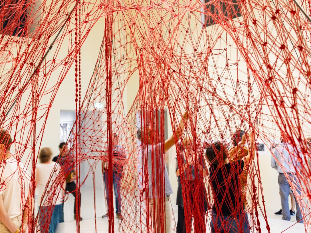 Verschiedene Personen knüpfen an einem raumhohen roten Netz in einem gelb gestrichenen Raum.
