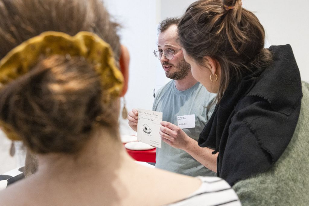 Zwei Frauen mit braunen hochgesteckten Haaren, die man nur von hinten sieht, schauen auf einen Mann im grauen T-Shirt mit einem weissen Namensschild, der ein weisses Blatt Papier vor seine Brust hält, auf dem etwas geschrieben und gezeichnet ist, das aber nicht erkennbar ist.