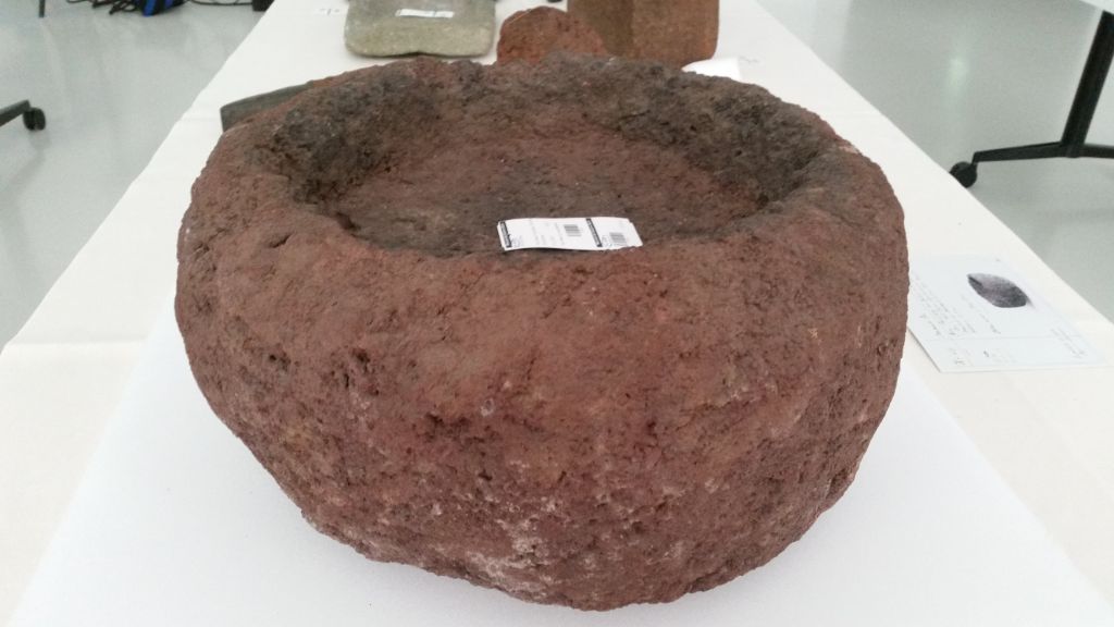 Auf einem Tisch steht eine Schale aus grobem rötlichen Stein. In der Schale liegt ein Zettel. Hinter der Schale liegen ein paar Dinge, die nicht erkennbar sind.