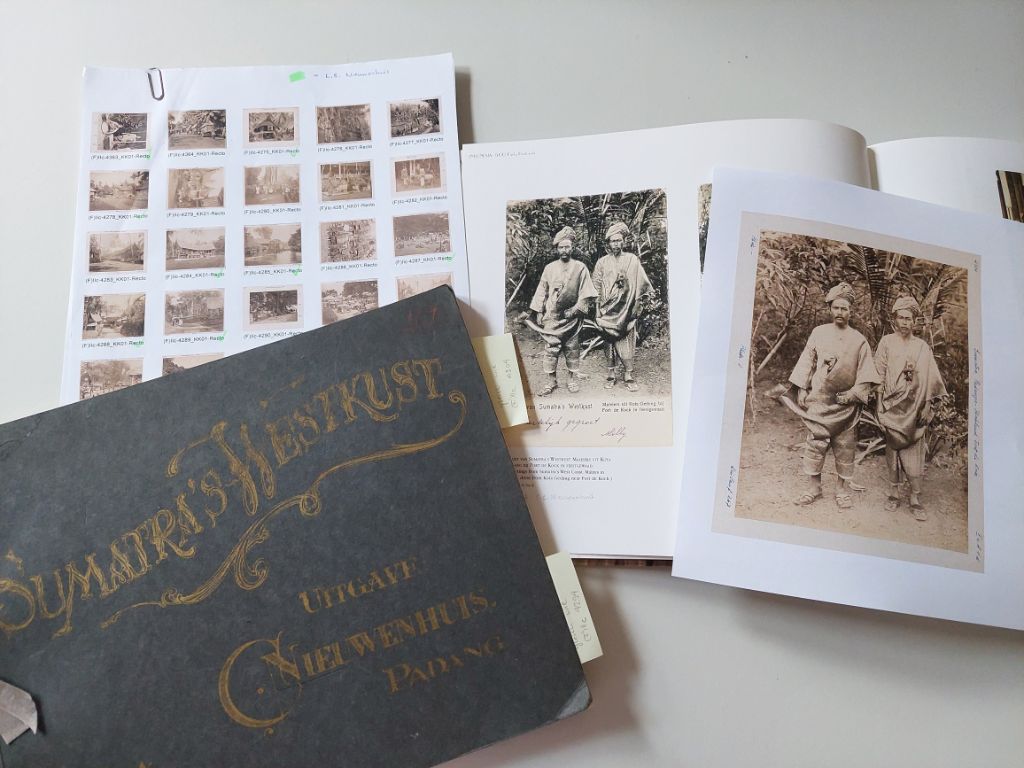 Auf grauem Hintergrund liegen 1 weisses Blatt, auf dem kleine Fotos fotokopiert sind und auf dem Blatt steckt noch eine Büroklammer. Darüber, das Blatt teilweise verdeckend liegt ein dunkelgraues Album mit goldenen Lettern mit dem Titel Sumatra's Westkust. Rechts davon ist ein Album aufgeschlagen, in dem Schwarz-Weiss-Fotos kleben und auf dem Album liegt eine Fotokopie eines Fotos von zwei Männern.