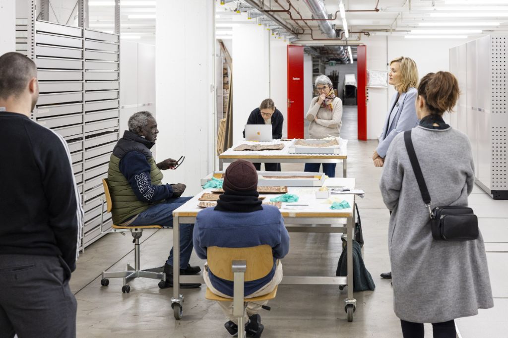 In einem hellen Kellerraum mit vielen Rollkorpussen stehen und sitzen Leute um zwei Tische. Zwei Männer und eine Frau diskutieren über Bilder, die auf den Tischen liegen. Zwei weitere Frauen hören zu. Eine Frau tippt in ein Laptop.