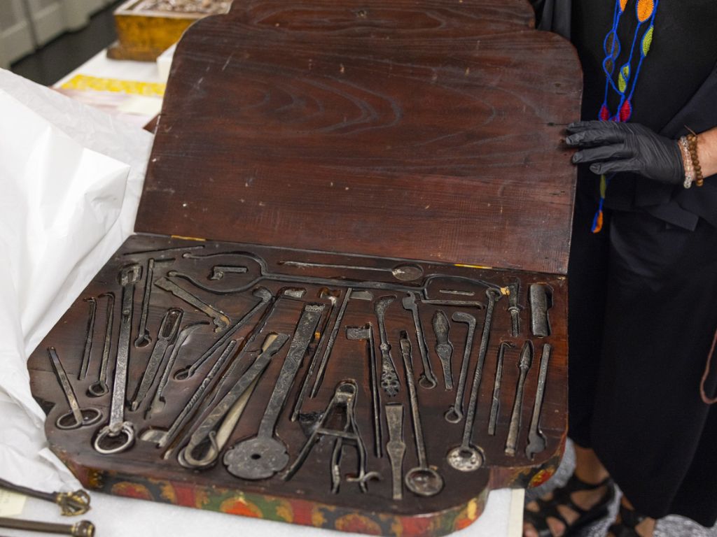 Eine Person, von der man fast nur eine Hand sieht, in einem schwarzen Handschuh, hält den Deckel einer rechteckigen Holzkiste auf, in der Instrumente liegen.