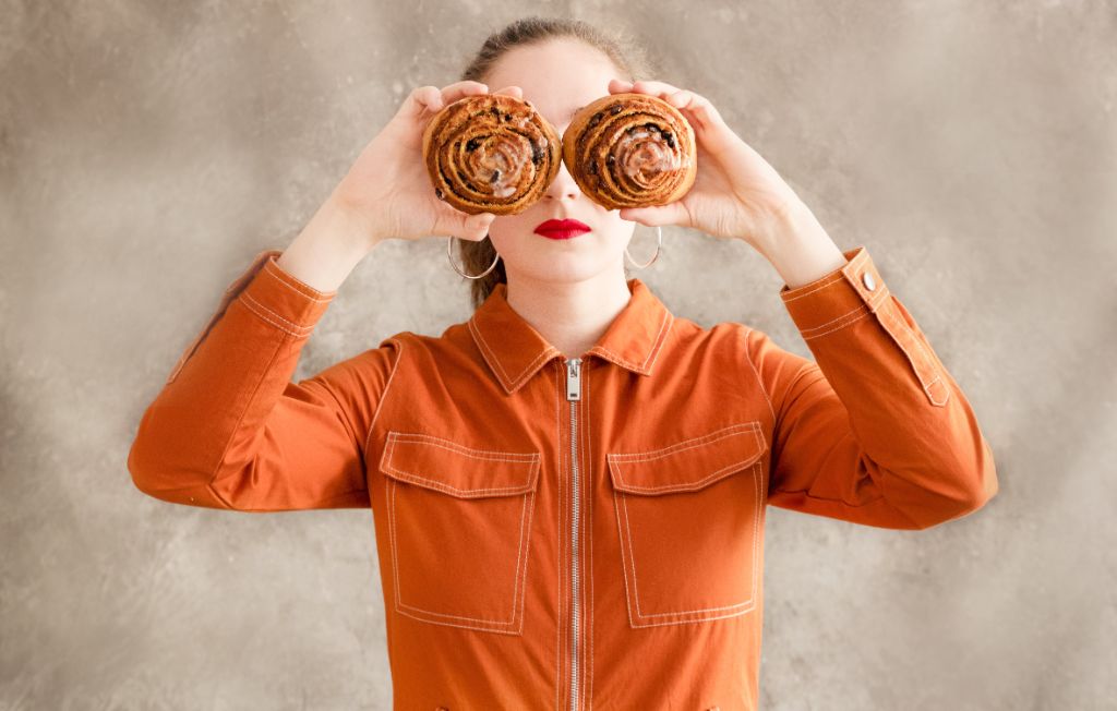 Eine Frau in einer orangen Jacke hält sich zwei Gebäckschnecken vor die Augen, so dass man von ihrem Gesicht nur die Stirne und den knallrot geschminkten Mund sieht.