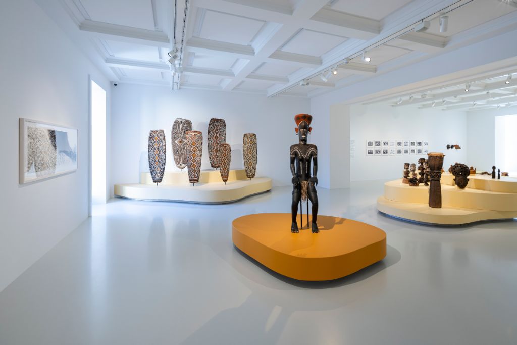 Blick in einen hellen grossen Ausstellungsraum, in dessen Mitte eine menschgrosse Figur auf einem orangen Podest steht. Links davon hängt ein Bild an der Wand, dahinter stehen auf einem gelben Podest sechs Schilde. Rechts von der Figur sind auf einem gelben Podest mehrere Figuren präsentiert, dahinter an der Wand Bilder, die man aber nicht erkennt.