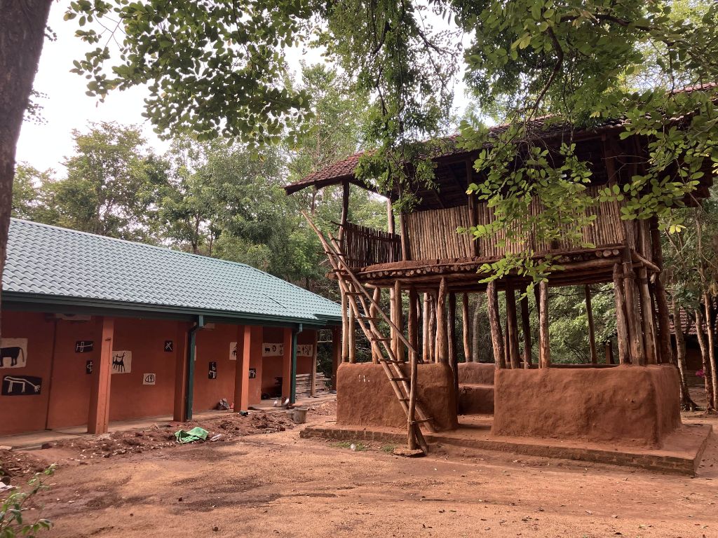 Zwei Gebäude umgeben von Laubbäumen. Links ein einstöckiges Haus mit grünem Ziegeldach und einer Veranda. Rechts ein zweistöckiges Holzhaus.