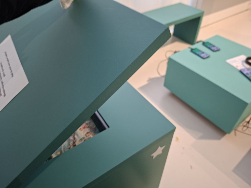 Bei einem türkisfarbenen, viereckigen Podest ist der Deckel etwas angehoben. Darin versteckt sich etwas Farbiges, das man aber nicht erkennen kann. Oben auf dem Deckel klebt ein weisser Zettel, auf dem ein paar schwarz geprintete Zeilen stehen, die aber ebenfalls kaum lesbar sind. An der Seite des Podests klebt ein weisser Stern. Im Hintergrund sieht man ein kleines türkisfarbenes Tischchen, auf dem Handys liegen.