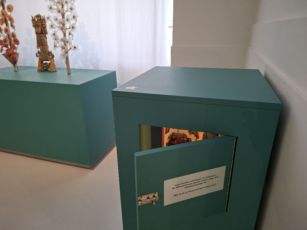 An einer weissen Wand steht ein viereckiger, türkisfarbener Kasten, auf dem ein weisser Stern klebt. Ein Türchen am Kasten ist etwas geöffnet und man erhascht einen Blick ins Innere, kann aber nicht wirklich sehen, was dort ist. Etwas leuchtet. Im Hintergrund ist ein Fenster mit einem weissen Vorhang und davor steht ein türkisfarbenes Podest auf dem zwei abstrakte Bäumchen stehen sowie eine Holzfigur.