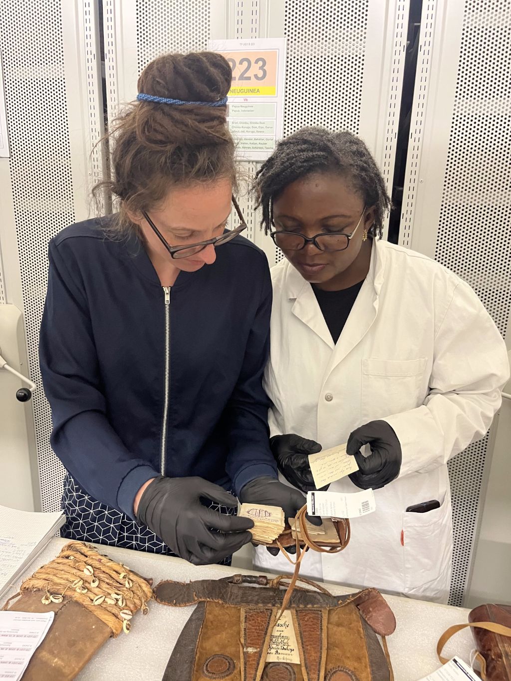 Vor Rollgestellen stehen zwei Frauen eng beieinander und schauen auf einen Gegenstand, den die Frau links in den Händen hält. Beide Frauen tragen schwarze Handschuhe. Vor ihnen steht ein Tisch, auf dem ein paar Gegenstände liegen.