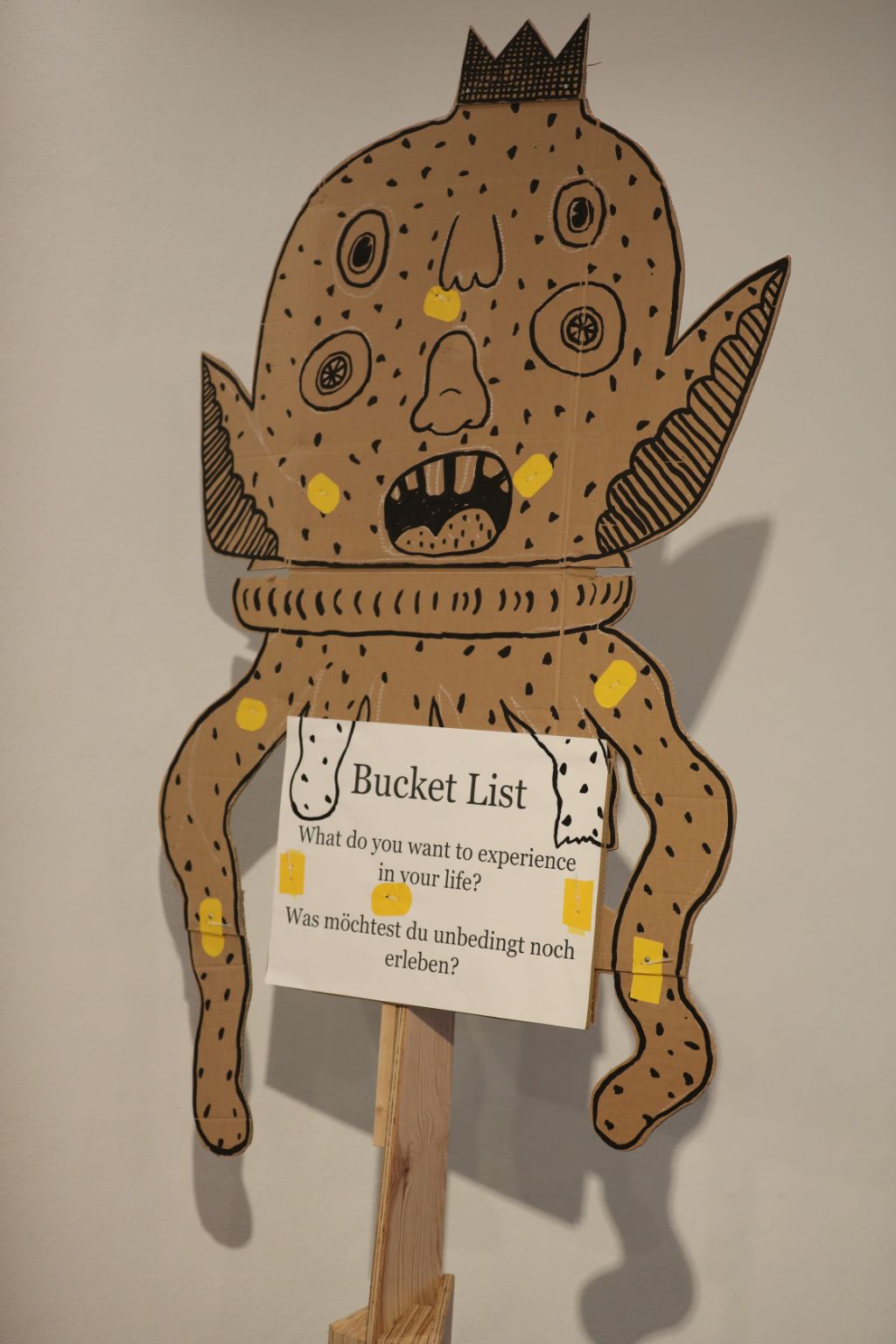 Auf einer Kartonfigur, die etwas einem Alien ähnelt, hängt ein Schild, auf dem steht: Bucket List. Was möchtest du unbedingt noch erleben?