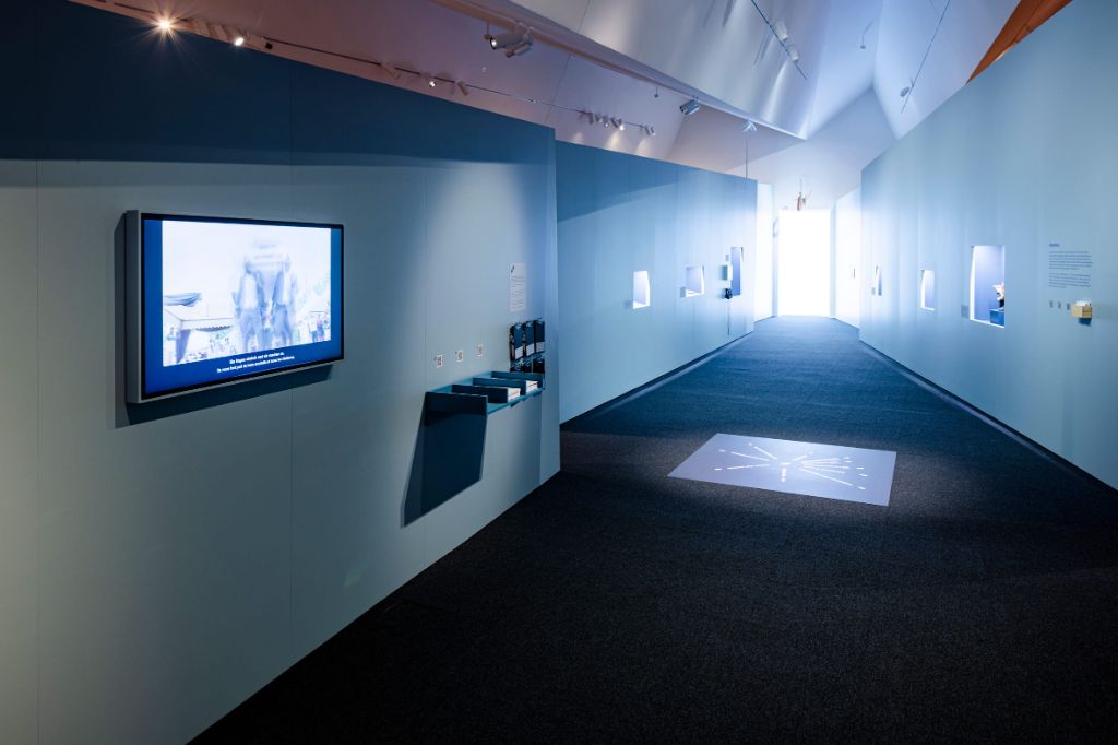 Ein langer Flur mit einem Bildschirm links an der Seitenwand, mehreren Nischen mit Objekten drin und einem raumhohen Fenster am Ende