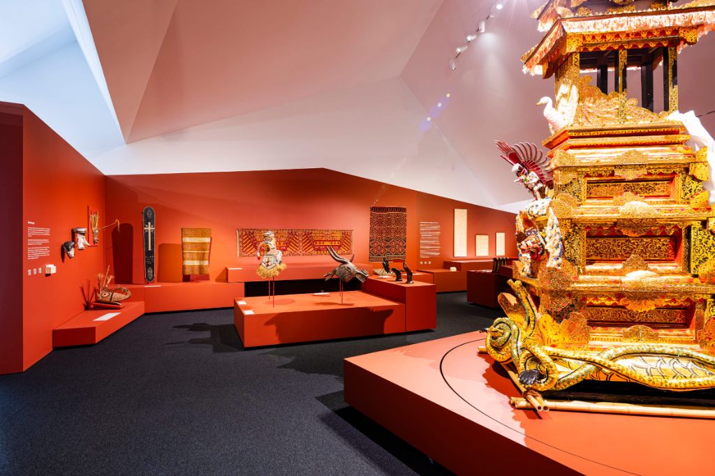 Ein Museumssaal mit roten Wänden zeigt goldene und bräunliche Objekte auf roten Podesten