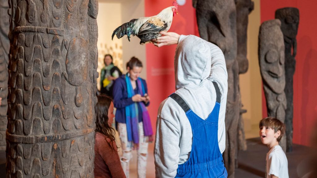 Ein Mann in einem blauen Overall und grauen Sweatshirt-Kapuzenpulli von hinten, auf seinem nach oben ausgestreckten Arm sitzt ein Huhn. Recht von ihm steht ein kleiner Junge und betrachtet mit geöffnetem Mund das Huhn. Im Hintergrund verschwommen zwei Frauen. Links vom Mann und im Hintergrund stehen grosse geschnitzte Hauspfosten vor roten Wänden.