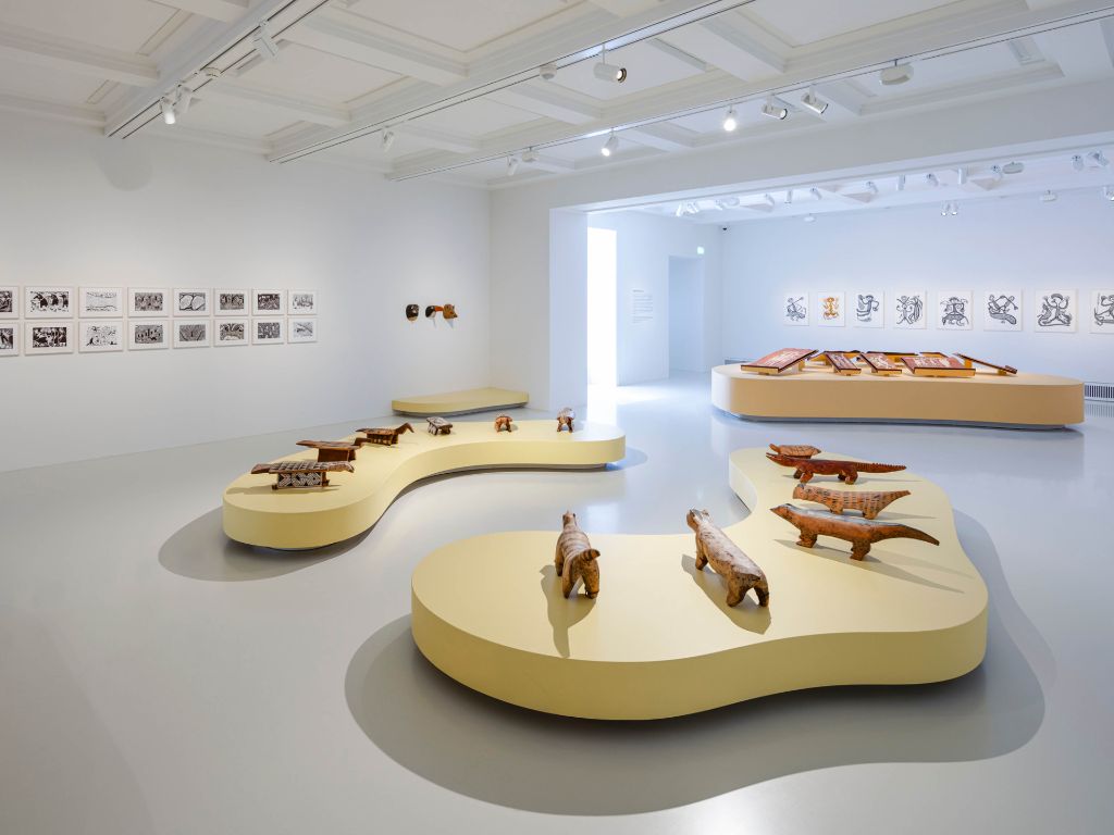 Ein Ausstellungsraum mit geschnitzten Tierfiguren auf kurvigen Podesten mit Kunstwerken an den Wänden.