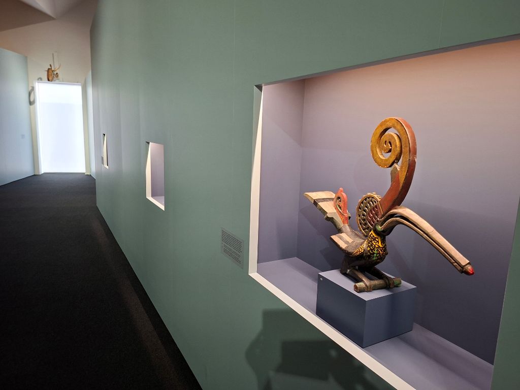 In einem grünen Gang, an dessen Ende ein grosses Fenster hell scheint, ist eine Nische zu sehen, in der auf einem kleinen blauen Podest die Figur eines Vogels aus Holz steht. Der Vogel hat einen sehr langen Schnabel und ein gerollte Horn auf dem Schnabel.