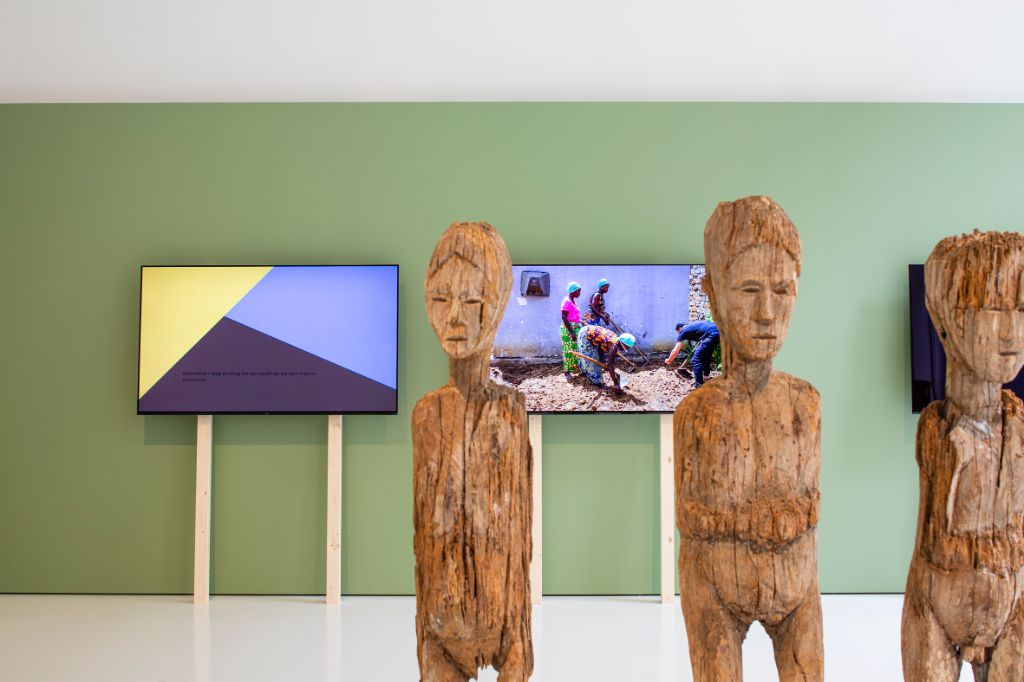 Drei Holzstatuen stehen vor drei Bildschirmen in einem Saal