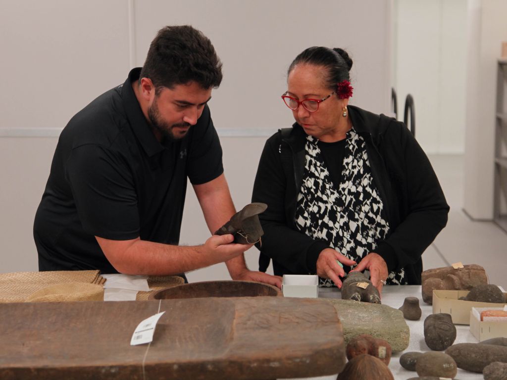 Zwei Personen betrachten an einem Tisch antike Objekte aus Holz und Stein in einem Innenraum.