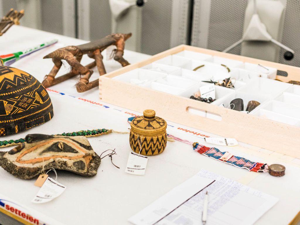 Tisch im Sammlungsdepot mit mehreren Objekten. Zu erkennen sind eine Tierfigur aus Holz, eine Maske aus Holz, und ein Perlenband.