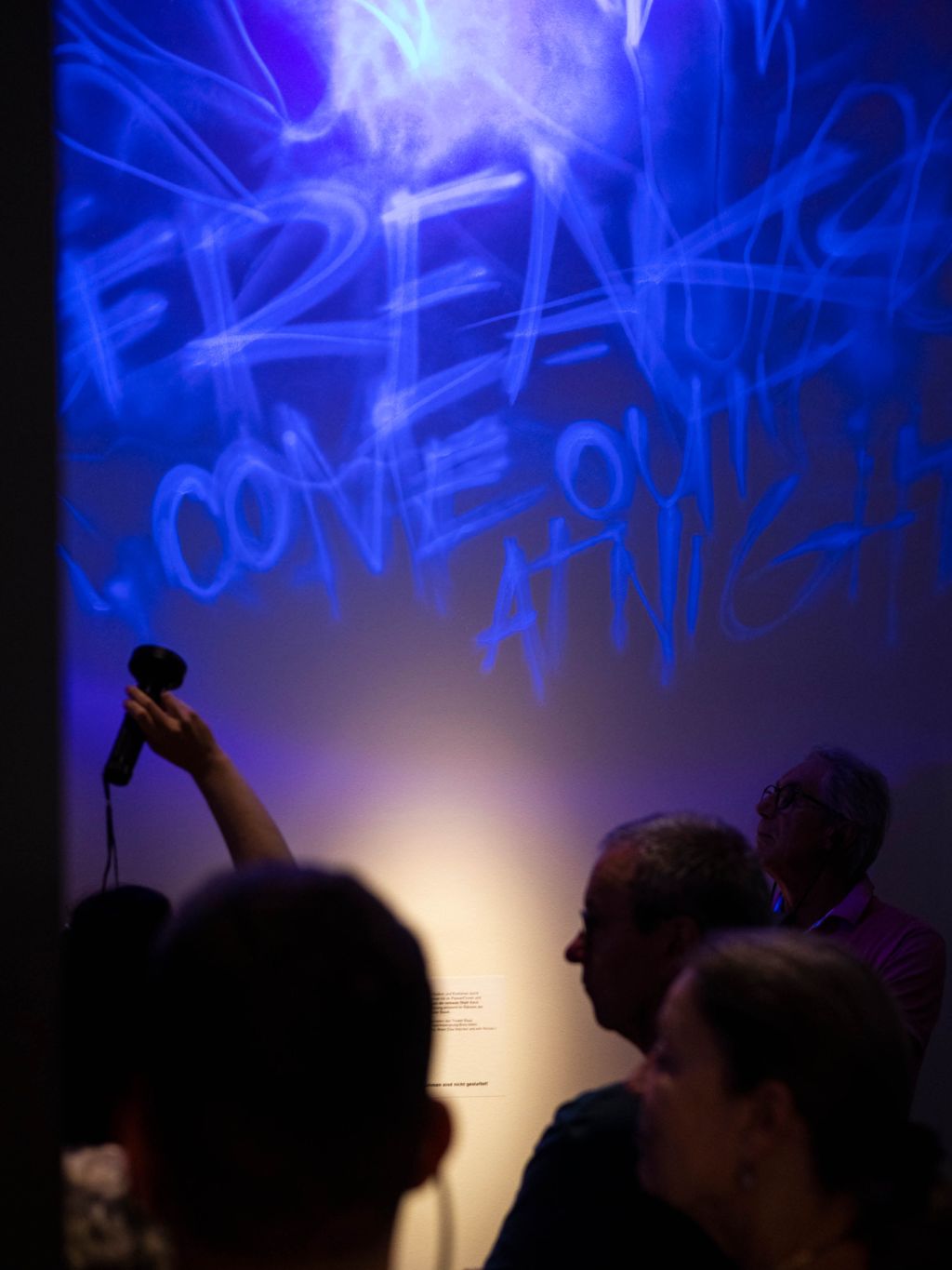 Wand in der Ausstellung Nacht mit neon-blauem Leuchtgraffiti, davor schattenhafte Umrisse von Besuchenden