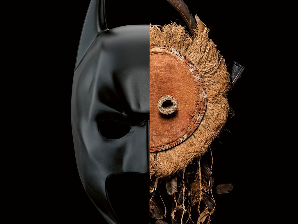 Zusammengesetzte Maske: links eine halbe Batman-Maske, rechts eine halbe, runde Maske aus braunen Naturfasern mit herabhängenden Vogelfedern